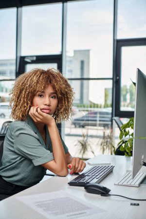 Téléchargez les photos : Jeune femme d'affaires afro-américaine bouclée assise au bureau devant un écran d'ordinateur au bureau - en image libre de droit