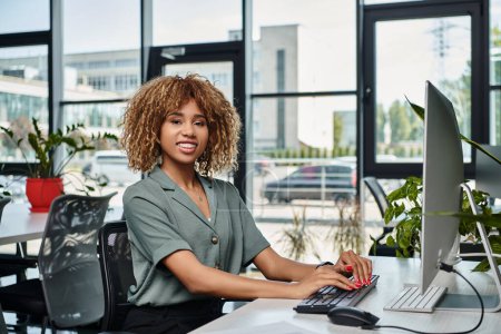 feliz joven mujer de negocios afroamericana sentada en el escritorio frente al monitor de la computadora en la oficina