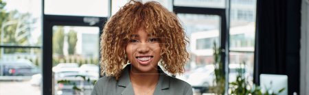 Téléchargez les photos : Bannière de heureuse femme d'affaires afro-américaine souriante et regardant la caméra dans le bureau contemporain - en image libre de droit