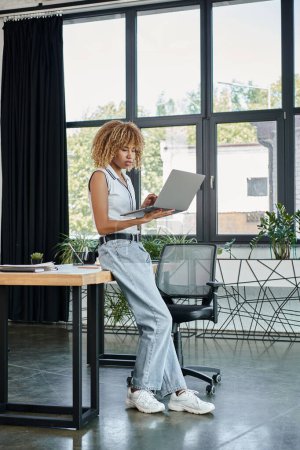 Téléchargez les photos : Femme d'affaires afro-américaine bouclée en tenue décontractée travaillant sur ordinateur portable dans une configuration de bureau moderne - en image libre de droit