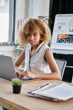 happy african american businesswoman in casual attire working on laptop at desk in modern office