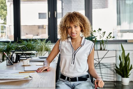 femme d'affaires afro-américaine joyeuse avec les cheveux bouclés assis à son bureau de travail avec papeterie