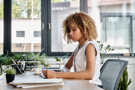 joven mujer de negocios afroamericana con el pelo rizado sentado en su mesa de trabajo y tomando notas