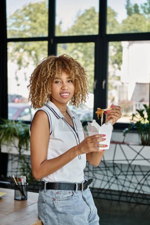 Téléchargez les photos : Heureux frisé afro-américain femme tenant repas à emporter dans la boîte en carton et en regardant la caméra - en image libre de droit