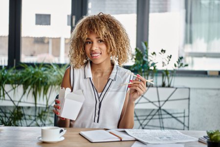 happy african american woman holding chopstick with noodles in carton box at desk, takeaway meal