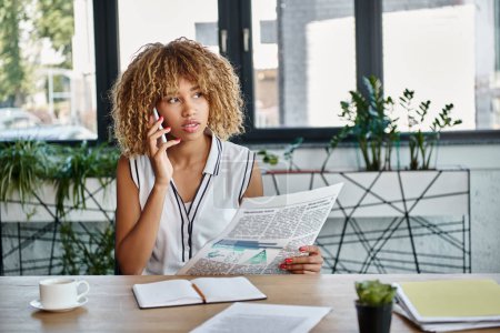 mujer de negocios afroamericana rizada hablando en un teléfono inteligente y sosteniendo el periódico en el escritorio en la oficina