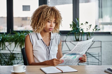 mujer de negocios afroamericana rizada leyendo el periódico y sentada en el escritorio en la oficina, profesión
