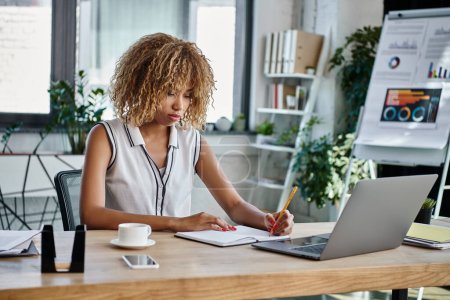 Dedicated african american businesswoman taking notes beside laptop and smartphone in office