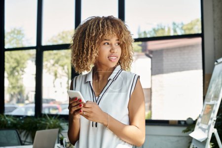 femme d'affaires afro-américaine bouclée et heureuse avec smartphone restant connecté au bureau ensoleillé