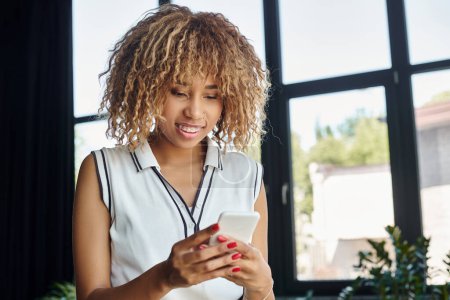 femme d'affaires afro-américaine bouclée et heureuse avec smartphone restant connecté au bureau ensoleillé