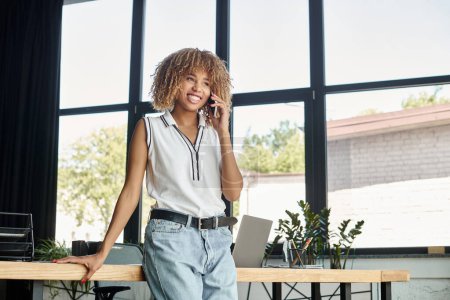 rizado y feliz afroamericana mujer de negocios teniendo llamada telefónica en la oficina moderna iluminada por el sol