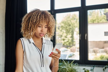 mujer de negocios afroamericana rizada y joven con teléfono inteligente permaneciendo conectado en la oficina iluminada por el sol