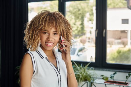 lockige und fröhliche afrikanisch-amerikanische Geschäftsfrau telefoniert im sonnendurchfluteten modernen Büro