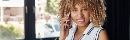 rizado y alegre mujer de negocios afroamericana que tiene llamada telefónica en la oficina moderna soleada, pancarta
