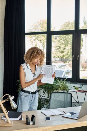jeune femme afro-américaine debout à son bureau et montrant contrat lors d'un appel vidéo sur ordinateur portable