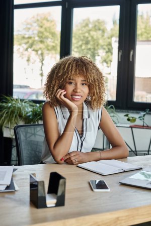 feliz joven mujer de negocios afroamericana sonriendo cerca de la computadora portátil y teléfono inteligente en el escritorio de trabajo
