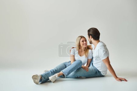 Téléchargez les photos : Un homme et une femme, incarnant l'amour et la proximité, assis sur le sol dans un studio gris - en image libre de droit