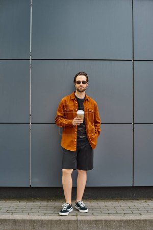A stylish man enjoying a cup of coffee while standing in front of a rustic brick wall.