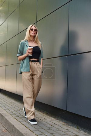Eine Frau in lässiger Kleidung steht neben einer Backsteinmauer, hält eine Tasse Kaffee in der Hand und nimmt sich einen Moment Zeit, um das Getränk zu genießen.
