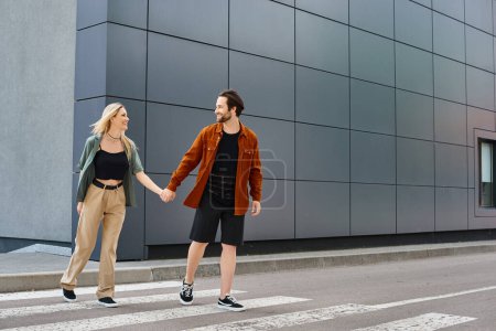 A sexy couple, a man and a woman, romantically walking across a street holding hands.