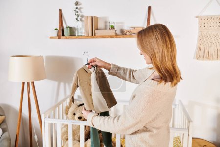 Téléchargez les photos : Jeune future mère tenant des vêtements de bébé près de la crèche avec des jouets mous dans la chambre maternelle, attente de naissance - en image libre de droit