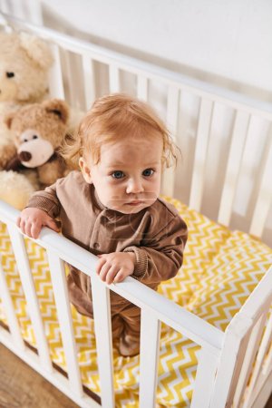 high angle view of toddler baby boy in romper standing in cozy crib in nursery room at home