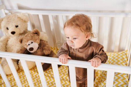 high angle view of little baby boy in romper standing in crib near soft toys in nursery room