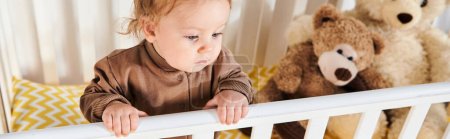 high angle view of toddler boy in romper standing in crib near soft toys in nursery room, banner