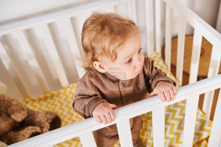 high angle view of cute little boy in romper standing in crib in nursery room at home, toddlerhood