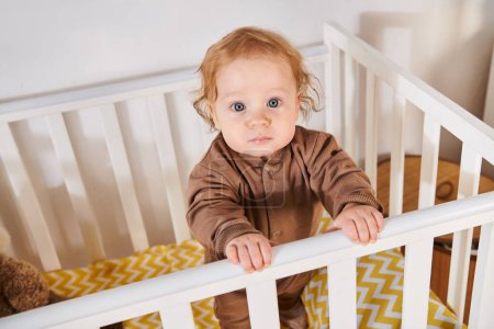 high angle view of adorable child in romper standing in crib in nursery room at home, toddlerhood