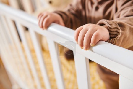 tiny hands of little boy standing in crib in nursery room at home, child growing in cozy apartment