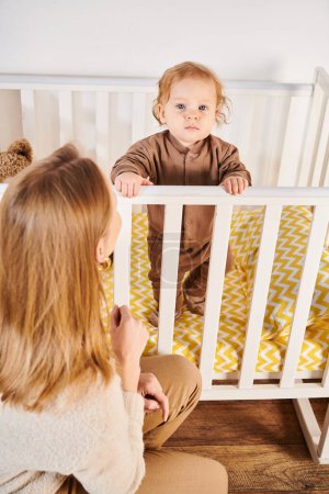 Photo for Woman looking at cute toddler son standing in crib in nursery room at home, happy motherhood - Royalty Free Image