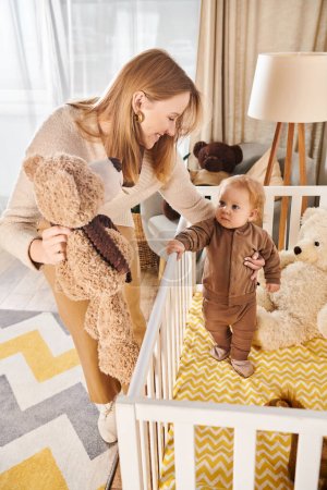 woman holding teddy bear near cute little son standing in crib in nursery room, happy motherhood