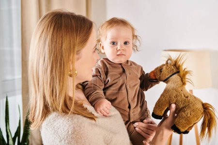 young woman holding cute son and toy horse in hands in nursery room at home, happy motherhood
