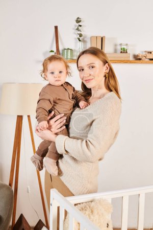 alegre mujer con adorable toddle hijo en manos mirando a cámara cerca cuna en cuarto de vivero en casa