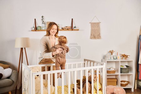 Photo for Young cheerful mother holding little toddler son near crib in nursery room, blissful motherhood - Royalty Free Image