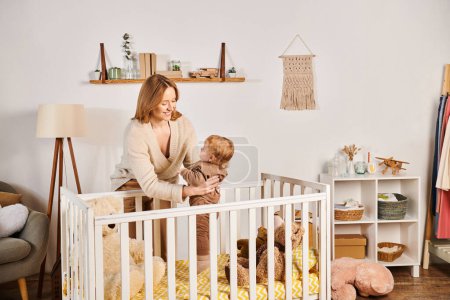 Photo for Joyful young mother holding cute toddler son near crib in nursery room, blissful motherhood - Royalty Free Image