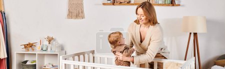 happy young mother holding cute toddle son near comfortable crib in nursery room, horizontal banner