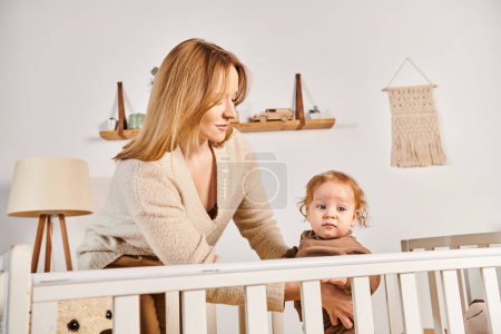Photo for Happy young mother supporting little child standing in crib in nursery room at home, motherhood - Royalty Free Image