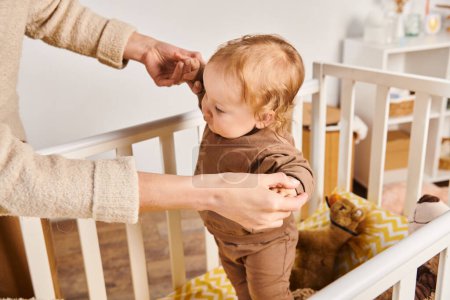 Photo for Young woman holding hands of little son standing in crib in nursery room at home, care and support - Royalty Free Image