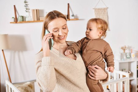 Photo for Smiling woman with little son in hands talking on mobile phone in nursery room, multitasking mother - Royalty Free Image