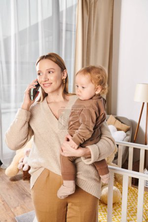happy woman with cute little son in hands talking on mobile phone at home, multitasking mother