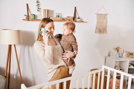 joyful woman with toddler kid in hands talking on mobile phone near crib, multitasking mother