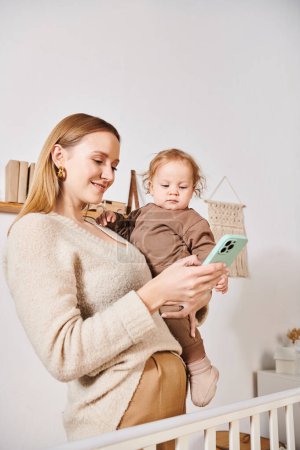 young mother with toddler son in hands messaging on mobile phone in nursery room, multitasking woman