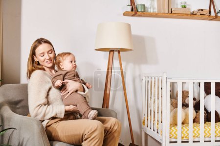 young woman with adorable son sitting in armchair near crib in nursery room, happy motherhood