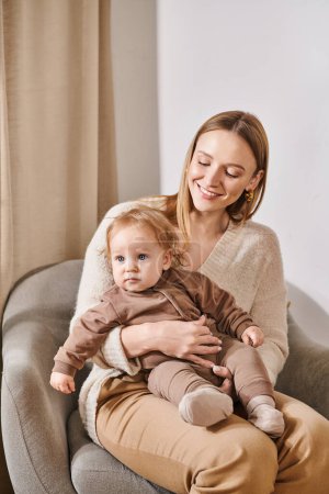 Photo for Cheerful attractive woman sitting in armchair with little boy in cozy nursery room, mother and son - Royalty Free Image
