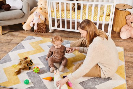 alegre madre e hijo pequeño jugando con juguetes blandos cerca de la cuna en la habitación de la guardería, crianza moderna