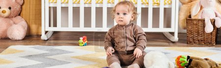niño pequeño sentado en el suelo cerca de juguetes blandos y cuna en la acogedora habitación del vivero, pancarta horizontal