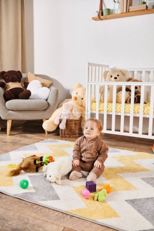 sweet little boy sitting on floor near soft toys and crib in cozy nursery room, happy toddlerhood