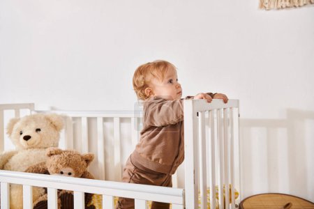 Neugieriger kleiner Junge im Kinderbett mit Plüschtieren im gemütlichen Kinderzimmer zu Hause, glückselige Kindheit